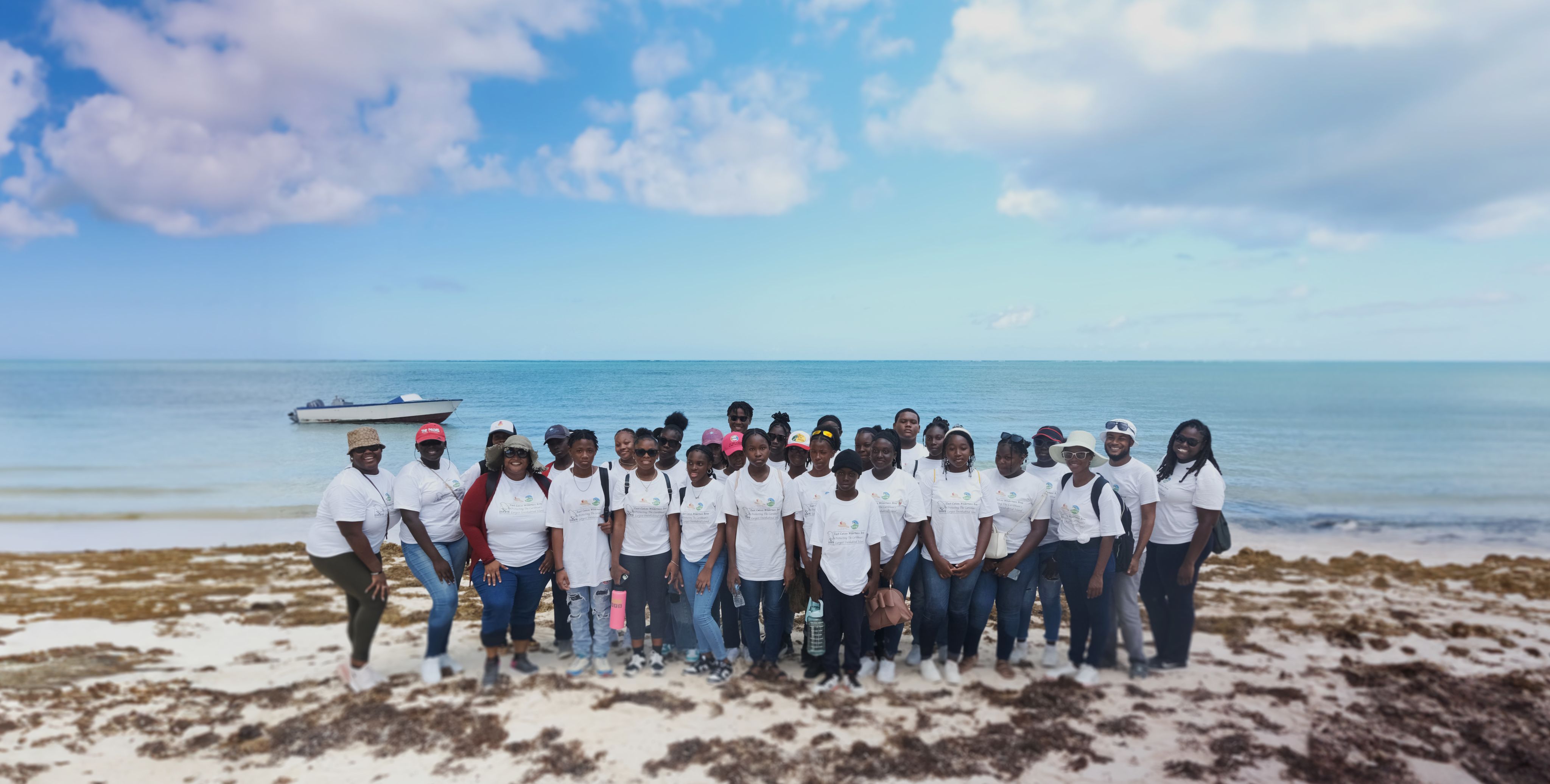 A group of schoolchildren standing on the sand covered with seaweed on East Caicos, with a small boat and the sea and sky in the background.